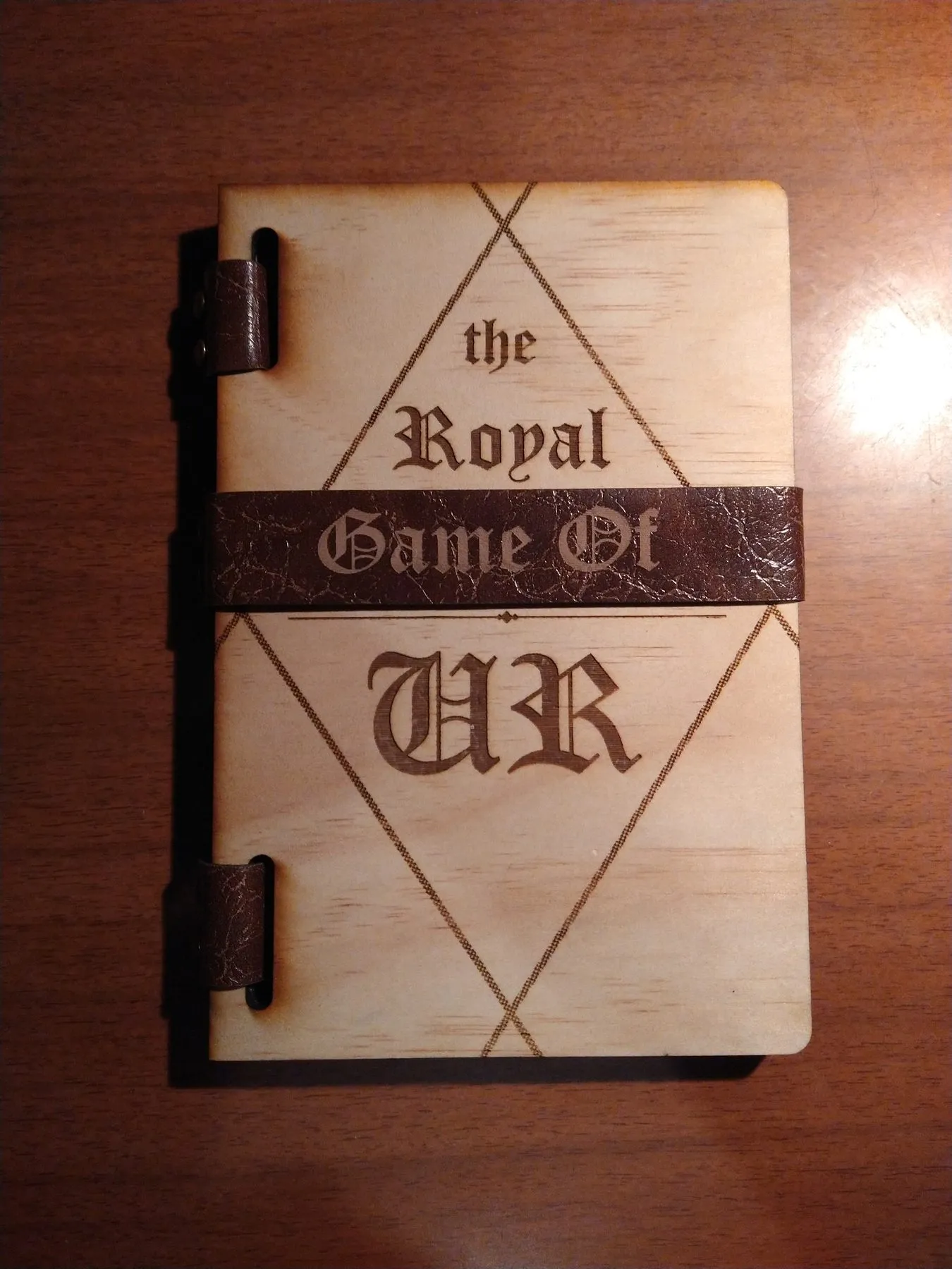 The Royal Game of Ur closed like a book, with the title engraved on a wooden cover and a faux-leather spine